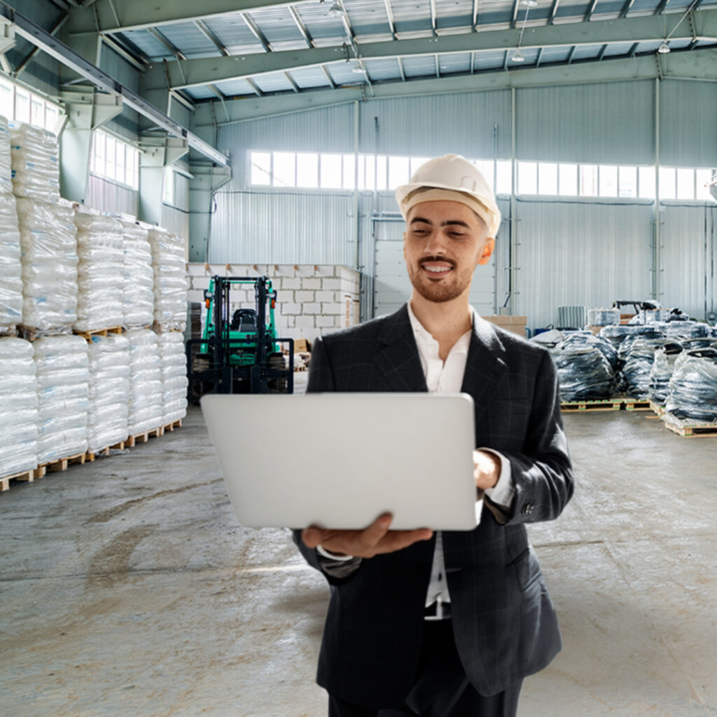 Imagem que representa o marketing industrial. Homem de negócios está em pé em uma ambiente que lembra uma fábrica, ele usa um capacete de proteção e carrega um notebook aberto em uma das mãos, enquanto digita com a outra.